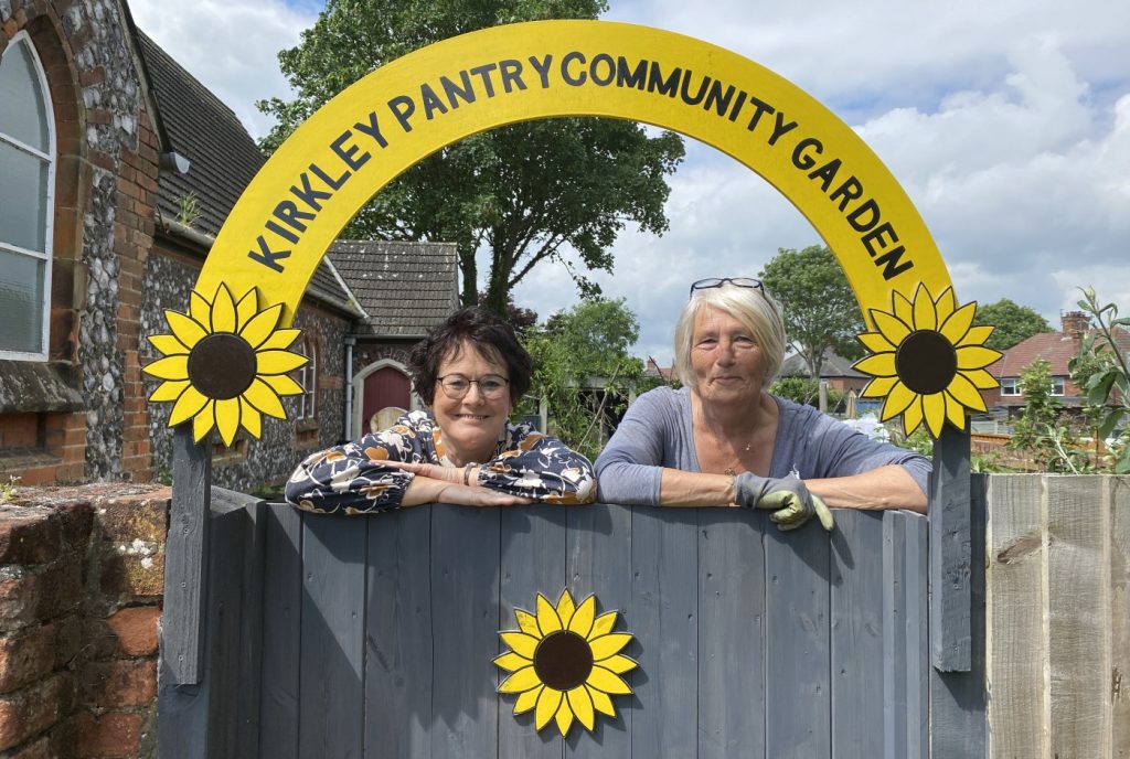 Paulette and Jackie lean on the gate, beneath a sign reading Kirkley Pantry Community Garden