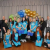 A large posed groupshot in a hall. Some people are in blue 'Your Local Pantry' aprons and there is a balloon arch behind them.