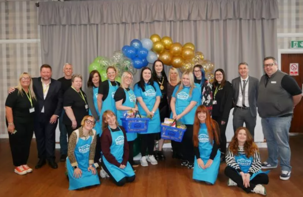 A large posed groupshot in a hall. Some people are in blue 'Your Local Pantry' aprons and there is a balloon arch behind them.