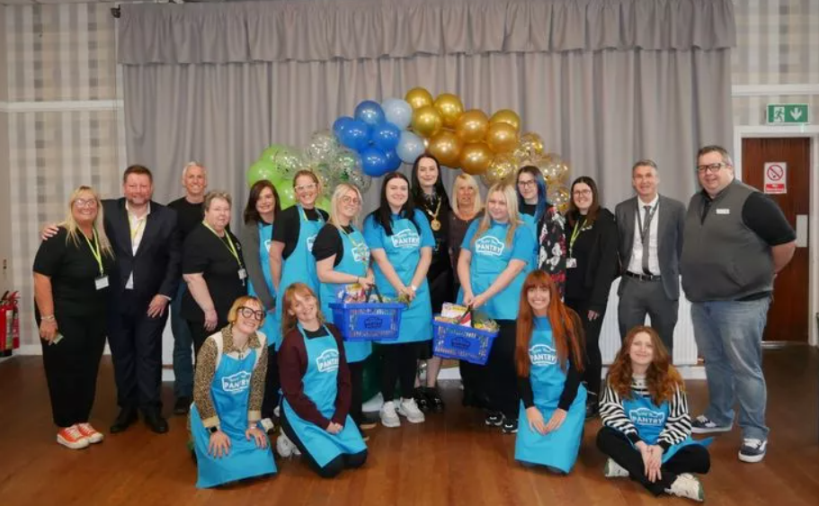 A large posed groupshot in a hall. Some people are in blue 'Your Local Pantry' aprons and there is a balloon arch behind them.