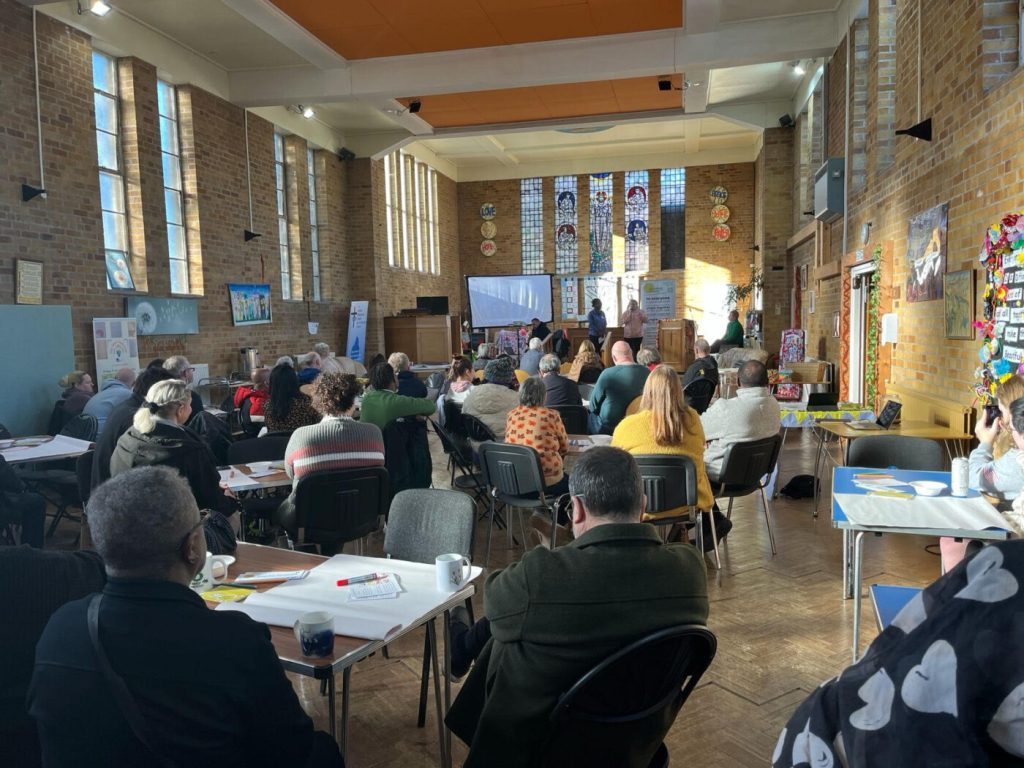 A view from the back of a busy church hall, with many people sitting and standing and chatting.