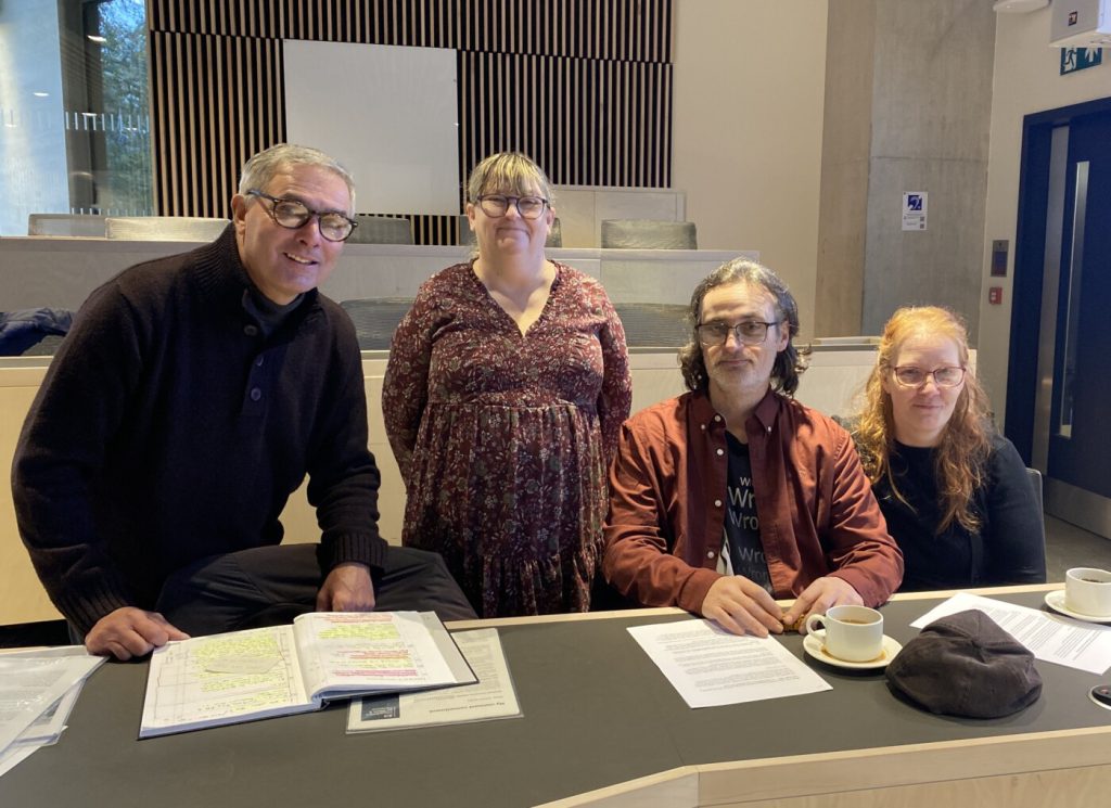 Four people behind a desk, looking at the camera. Two are standing; two sitting.