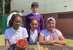 Four children sitting at a wooden picnic table outside a plain building.