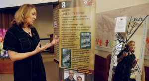 A woman stands beside a banner about homelessness, facing a mirror with words written on it.