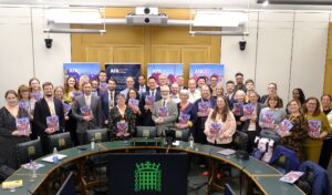 A large group of people behind a table at Westminster, with the Commons crest in the foreground