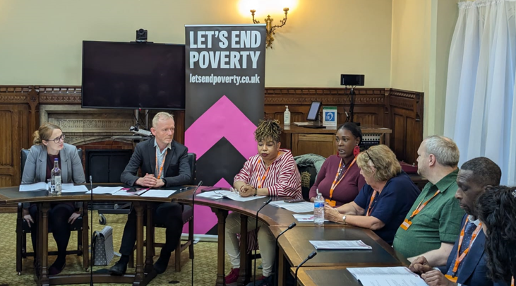 A row of people sitting behind a curving table, with a Let's End Poverty banner behind